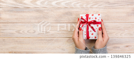 Woman hands give wrapped valentine or other holiday handmade present in paper with red ribbon. Present box, red heart decoration of gift on wooden table, top view with copy space for you design Woman hands give wrapped valentine or other holiday handmade present in paper with red ribbon. Present box, red heart decoration of gift on wooden table, top view with copy space for you design 139846325