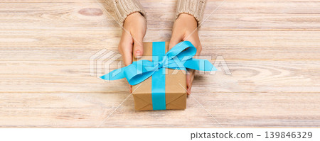 Woman's hands give wrapped valentine holiday handmade present in craft paper with blue ribbon. Present box, decoration of gift on black wooden table, POV with copy space Woman's hands give wrapped valentine holiday handmade present in craft paper with blue ribbon. Present box, decoration of gift on black wooden table, POV with copy space 139846329