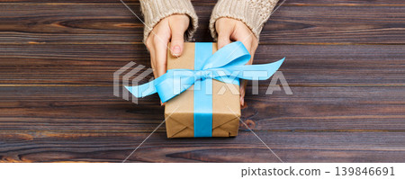 Woman's hands give wrapped valentine holiday handmade present in craft paper with blue ribbon. Present box, decoration of gift on black wooden table, POV with copy space Woman's hands give wrapped valentine holiday handmade present in craft paper with blue ribbon. Present box, decoration of gift on black wooden table, POV with copy space 139846691