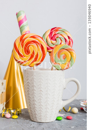 Colorful candies in cup on table on light background background. Large swirled lollipops. Creative concept of a jar full of delicious sweets from the candy store Colorful candies in cup on table on light background background. Large swirled lollipops. Creative concept of a jar full of delicious sweets from the candy store 139846940