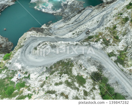 Aerial view of limestone quarry with heavy truck moving along winding road during mining operations, highlighting raw material supply, industrial activity, and environmental impact on landscape. Aerial view of limestone quarry with heavy truck moving along winding road during mining operations, highlighting raw material supply, industrial activity, and environmental impact on landscape. 139847282
