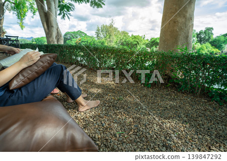Barefoot woman enjoying digital detox by reading book outdoors at riverside hotel resort with natural stone ground and cool breeze for peaceful self-reflection and mental wellness escape. Self-care. 139847292