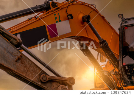 Closeup hydraulic piston arm of dirty backhoe at construction site. Yellow excavator parked at construction site against sunset sky. Digger machine. Hydraulic machinery. Excavator for rent business. Closeup hydraulic piston arm of dirty backhoe at construction site. Yellow excavator parked at construction site against sunset sky. Digger machine. Hydraulic machinery. Excavator for rent business. 139847624