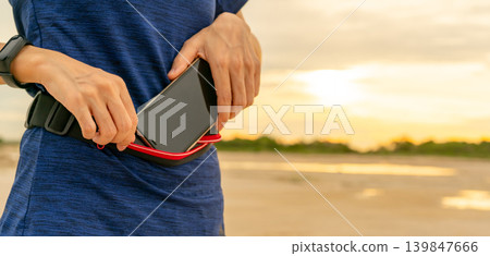 Young Asian woman keep smartphone in waist bag before running cardio exercise in the morning. Outdoor workout. Runner and smart band wearable device. Gadget for healthy lifestyle. Jogging fashion. Young Asian woman keep smartphone in waist bag before running cardio exercise in the morning. Outdoor workout. Runner and smart band wearable device. Gadget for healthy lifestyle. Jogging fashion. 139847666