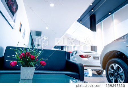 Fake red flowers in vase on table in modern car showroom. Car dealership interior building. Artificial flower on blurred background of new white luxury car parked in showroom. Automotive industry. Fake red flowers in vase on table in modern car showroom. Car dealership interior building. Artificial flower on blurred background of new white luxury car parked in showroom. Automotive industry. 139847830