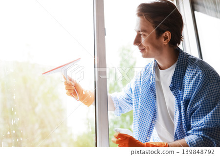 Smiling young man cleaning a window with a squeegee and spray bottle, wearing protective rubber gloves during springtime house cleaning chores. Smiling young man cleaning a window with a squeegee and spray bottle, wearing protective rubber gloves during springtime house cleaning chores. 139848792
