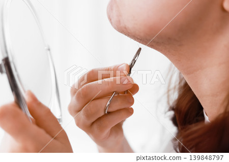 Woman looking in a mirror during her home beauty routine, using a tool for facial hair removal on her chin to manage unwanted hair caused by hormonal changes. Woman looking in a mirror during her home beauty routine, using a tool for facial hair removal on her chin to manage unwanted hair caused by hormonal changes. 139848797
