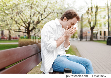 Man suffering from spring allergies, sneezing into his hands while sitting on a park bench surrounded by blooming trees. Seasonal hay fever concept. 139848870