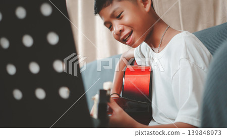 Happy Asian boy singing and playing acoustic guitar on sofa at home. Child musician with joyful expression practicing music. Lifestyle, hobby and education concept. Happy Asian boy singing and playing acoustic guitar on sofa at home. Child musician with joyful expression practicing music. Lifestyle, hobby and education concept. 139848973