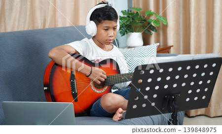 Asian boy wearing headphones playing acoustic guitar and looking at music sheet while learning musical instrument online via laptop in living room at home. Asian boy wearing headphones playing acoustic guitar and looking at music sheet while learning musical instrument online via laptop in living room at home. 139848975