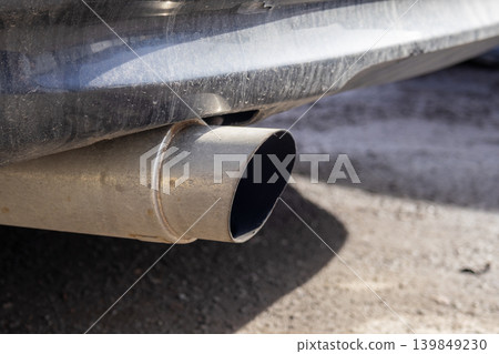 The car exhaust pipe is visible at the back of the vehicle in a parking lot. Sunlight shines on the metal surface and surrounding ground The car exhaust pipe is visible at the back of the vehicle in a parking lot. Sunlight shines on the metal surface and surrounding ground 139849230