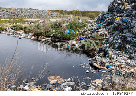Garbage is piled up around a small pond with some plants nearby in a landfill area under a cloudy sky Garbage is piled up around a small pond with some plants nearby in a landfill area under a cloudy sky 139849277