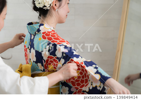 An image of a Japanese woman checking her appearance in a yukata in a mirror, with a person preparing to put it on and the woman wearing the yukata, showing only her back and no face. An image of a Japanese woman checking her appearance in a yukata in a mirror, with a person preparing to put it on and the woman wearing the yukata, showing only her back and no face. 139849414