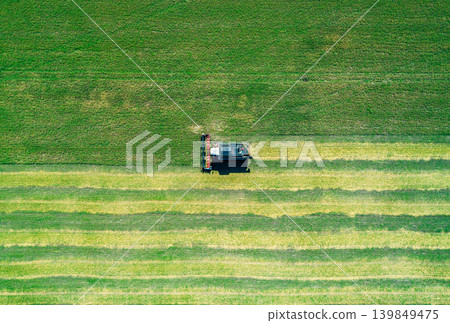 Top down view shows a large combine harvester cuts rows of green crops in a sprawling farm field under a bright blue sky Top down view shows a large combine harvester cuts rows of green crops in a sprawling farm field under a bright blue sky 139849475
