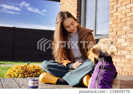 A happy young woman sitting cross-legged outdoors with her small dog on a sunny day, cozy autumn outfit and calm lifestyle scene, pet human friendship A happy young woman sitting cross-legged outdoors with her small dog on a sunny day, cozy autumn outfit and calm lifestyle scene, pet human friendship 139849776