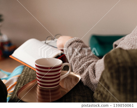 Unrecognizable woman in grey bathrobe relaxing with a book and hot drink on a green corduroy sofa at home. A middle-aged woman relaxing indoors. Unrecognizable woman in grey bathrobe relaxing with a book and hot drink on a green corduroy sofa at home. A middle-aged woman relaxing indoors. 139850061