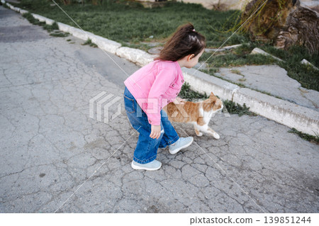 Little Caucasian girl walking with a ginger cat on a residential street while showing affection and care for domestic animals. Childhood lifestyle Little Caucasian girl walking with a ginger cat on a residential street while showing affection and care for domestic animals. Childhood lifestyle 139851244