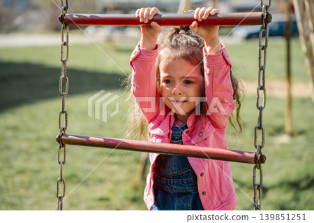 Little Caucasian girl with a worried facial expression climbing rope playground bars while wearing a pink jacket outdoors. Childhood lifestyle 139851251