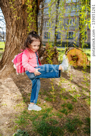 Little Caucasian girl playing near a tree with hanging green branches while wearing a pink jacket and denim clothes outdoors. Childhood lifestyle 139851261