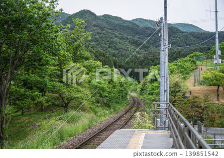 A single-track railway line stretching towards the lush green mountains, viewed from the platform of an unmanned station in early summer. 139851542