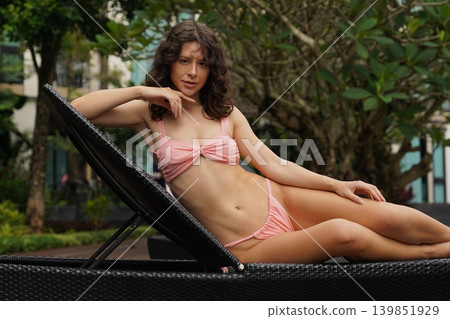 A woman in a brightly colored swimsuit poses on a lounge chair by the pool A woman in a brightly colored swimsuit poses on a lounge chair by the pool 139851929