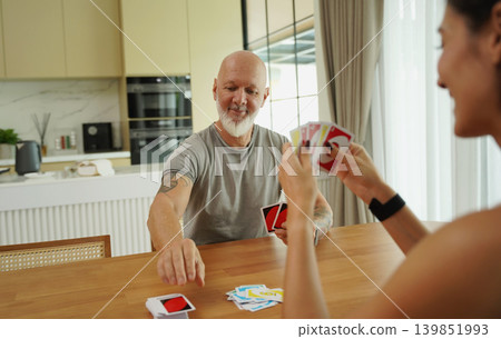 A man and a woman play cards at a wooden table in a sunlit modern living room A man and a woman play cards at a wooden table in a sunlit modern living room 139851993