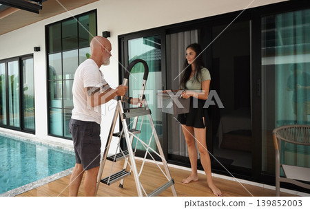Two individuals collaborate carefully to mount outdoor lighting near their modern home Two individuals collaborate carefully to mount outdoor lighting near their modern home 139852003
