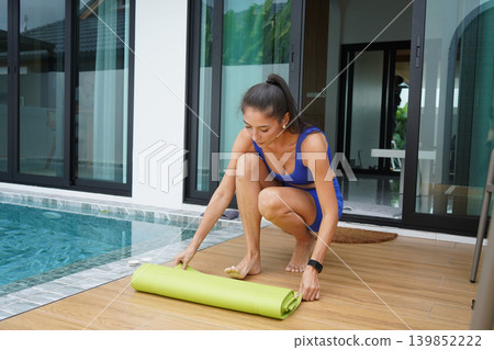 woman rolling yoga mat by pool preparing for morning stretch on deck wearing blue shorts lace woman rolling yoga mat by pool preparing for morning stretch on deck wearing blue shorts lace 139852222