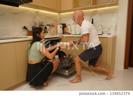 Joyful young couple sorts and loads kitchen utensils into dishwasher with warm ambiance Joyful young couple sorts and loads kitchen utensils into dishwasher with warm ambiance 139852379