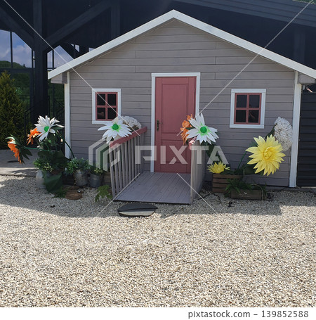 A fairy-tale wooden house with a pink door and large artificial flowers in the yard. A cute decorative location A fairy-tale wooden house with a pink door and large artificial flowers in the yard. A cute decorative location 139852588