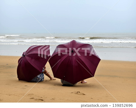 Two travelers sheltered under deep red umbrellas gazing at ocean Two travelers sheltered under deep red umbrellas gazing at ocean 139852696