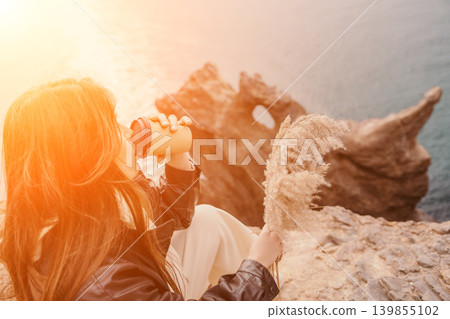 Woman coffee cliff drinking from paper cup while sitting on rocky edge overlooking ocean during warm sunset travel lifestyle scene Woman coffee cliff drinking from paper cup while sitting on rocky edge overlooking ocean during warm sunset travel lifestyle scene 139855102
