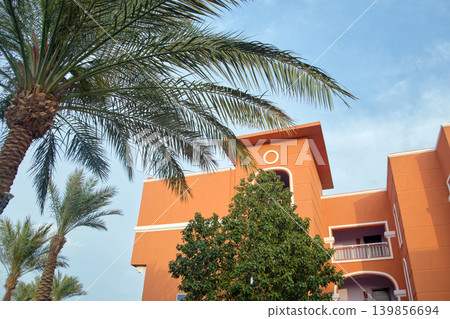Fragment of red brick resort hotel exterior with palm trees under blue summer sky Fragment of red brick resort hotel exterior with palm trees under blue summer sky 139856694