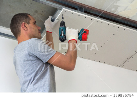 Construction worker assemble a suspended ceiling with drywall and fixing the drywall to the ceiling metal frame with screwdriver. Construction worker assemble a suspended ceiling with drywall and fixing the drywall to the ceiling metal frame with screwdriver. 139856794