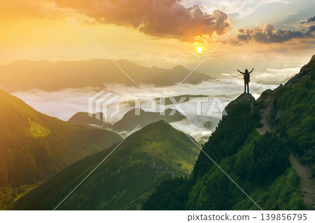 Wide mountain panorama. Small silhouette of tourist with backpack on rocky mountain slope with raised hands over valley covered with white puffy clouds. Beauty of nature, tourism and traveling concept Wide mountain panorama. Small silhouette of tourist with backpack on rocky mountain slope with raised hands over valley covered with white puffy clouds. Beauty of nature, tourism and traveling concept 139856795