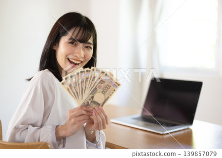 A young Japanese woman smiles in front of a computer desk, holding ten 10,000 yen bills. 139857079