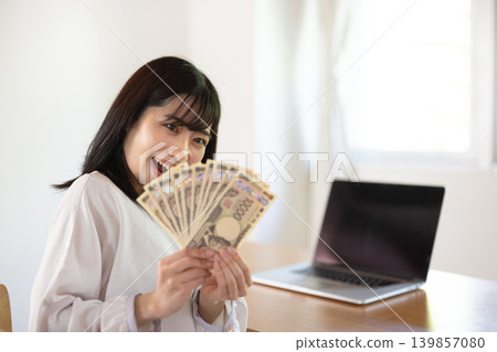 A young Japanese woman smiles in front of a computer desk, holding ten 10,000 yen bills. A young Japanese woman smiles in front of a computer desk, holding ten 10,000 yen bills. 139857080