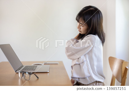 A young Japanese woman with her hand on her shoulder and frowning while working on her computer. A young Japanese woman with her hand on her shoulder and frowning while working on her computer. 139857251