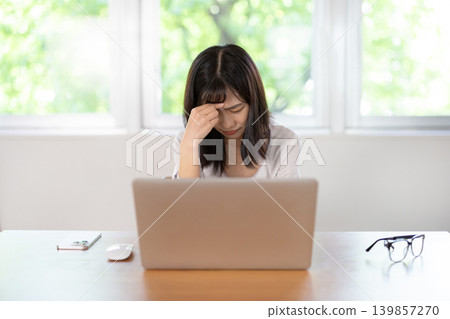一位年輕的日本女子一邊用手扶著頭，一邊皺著眉頭使用電腦。 139857270