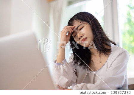 A young Japanese woman puts her hand to her head and frowns while working on her computer. A young Japanese woman puts her hand to her head and frowns while working on her computer. 139857273
