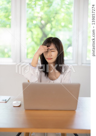 A young Japanese woman puts her hand to her head and frowns while working on her computer. A young Japanese woman puts her hand to her head and frowns while working on her computer. 139857274
