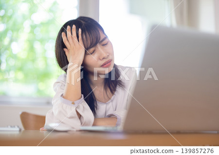 一位年輕的日本女子一邊用手扶著頭，一邊皺著眉頭使用電腦。 139857276