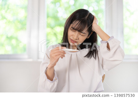 A young Japanese woman in her living room looks distressed while looking at a thermometer. A young Japanese woman in her living room looks distressed while looking at a thermometer. 139857530