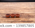 Three handcrafted terracotta clay tea cups placed side by side on an aged rustic wooden table with one cup filled with dark tea and two empty cups in warm earthy tones against a dark background 139857805