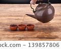 Hand holding antique brown ceramic teapot with brass handle pouring hot tea into three small terracotta clay cups arranged in a row on a rustic weathered wooden table surface 139857806