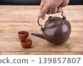 Hand Holding Antique Brown Ceramic Teapot Pouring Golden Tea Into Traditional Small Clay Cups on Weathered Rustic Wooden Table Surface With Dark Background in Studio Setting 139857807