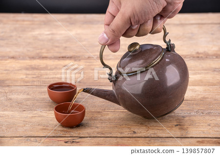 Hand Holding Antique Brown Ceramic Teapot Pouring Golden Tea Into Traditional Small Clay Cups on Weathered Rustic Wooden Table Surface With Dark Background in Studio Setting Hand Holding Antique Brown Ceramic Teapot Pouring Golden Tea Into Traditional Small Clay Cups on Weathered Rustic Wooden Table Surface With Dark Background in Studio Setting 139857807
