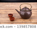 Traditional antique brown ceramic teapot with ornate brass handle accompanied by two small terracotta clay tea cups on a weathered rustic wooden table against a dark background in warm tones 139857808