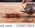 Hand Holding an Antique Brown Teapot Pouring Fresh Hot Tea Into Three Small Traditional Terracotta Clay Cups Arranged in a Row on a Rustic Weathered Wooden Table with Dark Background 139857809