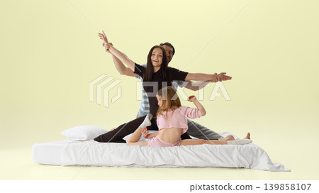 Playful family posing like dancers on white bed in pastel yellow studio. 139858107